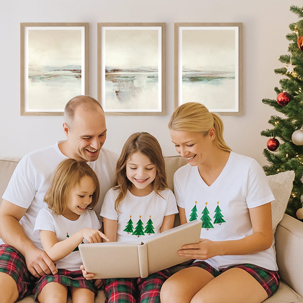 Happy family sitting on a couch, looking at a photo album, all wearing matching Christmas pyjamas, in a living room decorated for Christmas and wall art poster frames on the wall.